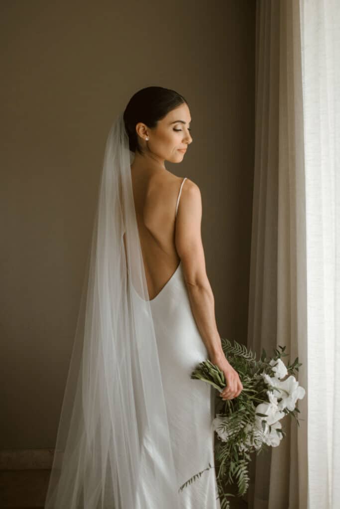 Bride wearing a minimalist satin wedding gown and long veil, holding a white floral bouquet beside a window.