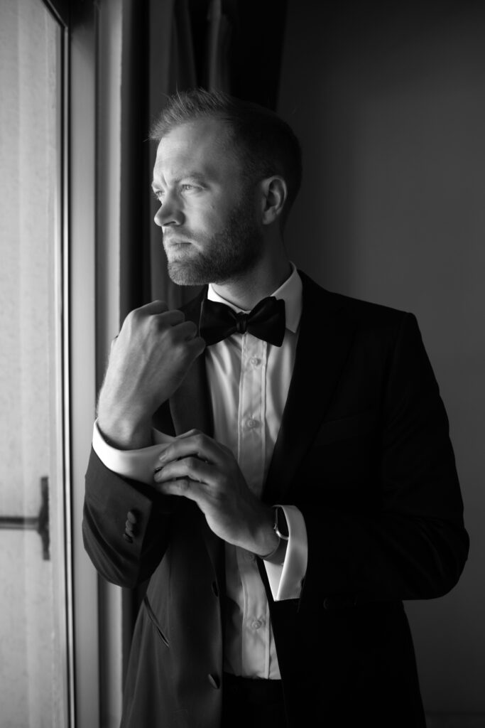 Groom adjusting his black bow tie beside a window before his elegant destination wedding in Cabo.