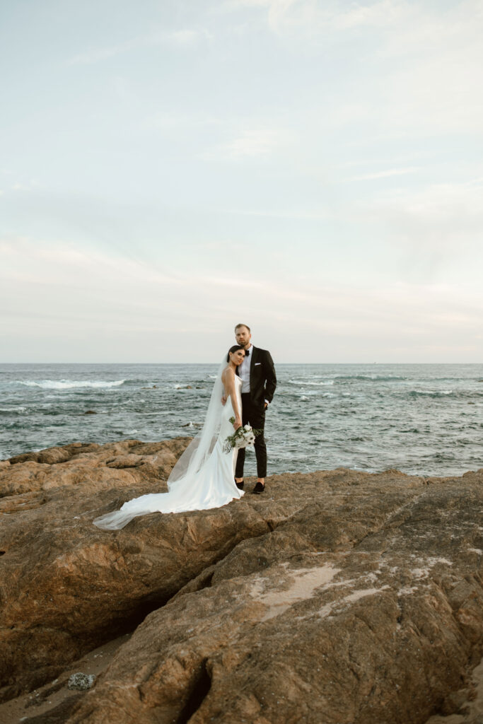 Christine and Alex posing together on coastal rocks overlooking the ocean during their Los Cabos destination wedding.