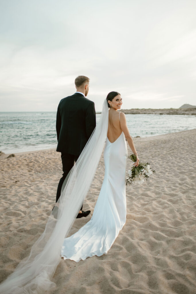 Bride smiling over her shoulder while walking along a sandy Cabo beach with her groom and flowing veil.