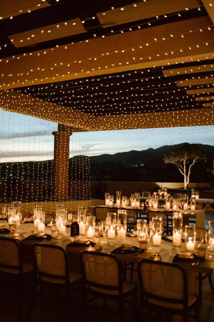 Outdoor wedding reception with long dining tables, glowing candles and cascading string lights at sunset in Los Cabos.