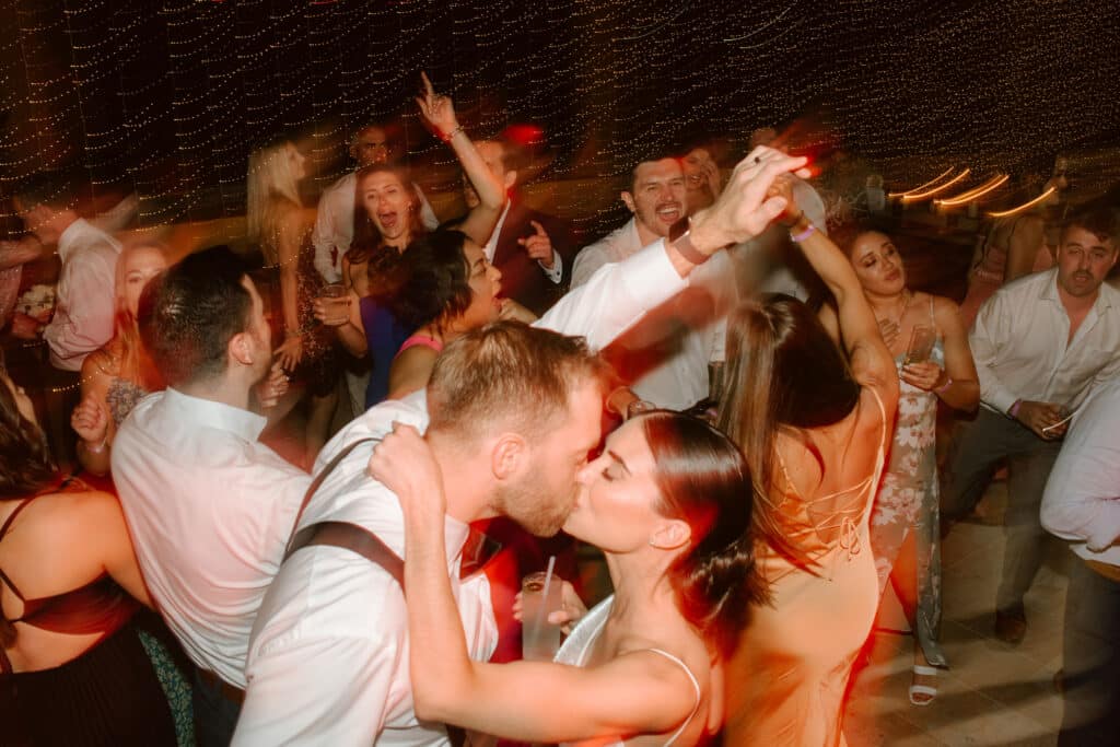 Christine and Alex kissing on the dance floor surrounded by celebrating guests beneath twinkling lights at their Cabo wedding.
